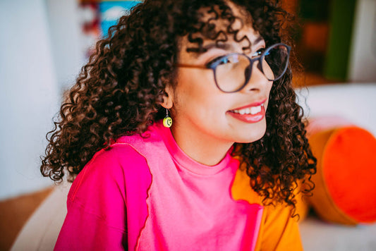 Smiling woman with curly hair and glasses wearing a pink and orange shirt, seated indoors with a cheerful vibe
