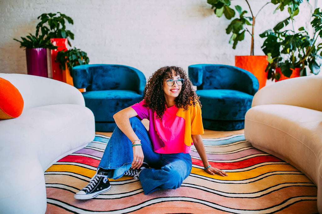 Teen girl with curly hair and glasses sitting on a colorful striped rug, surrounded by vibrant furniture and plants, smiling confidently